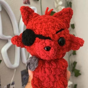May include: A red crocheted plush toy shaped like a fox with pointed ears, a black eye patch over one eye, a small black nose, and a grey hook-shaped accessory attached to its side. The plush is crafted from textured yarn with a soft appearance.