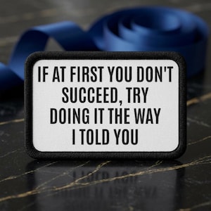 Puede incluir: Un parche rectangular bordado con fondo blanco y un borde negro grueso. El parche presenta texto bordado en negro que dice: IF AT FIRST YOU DON'T SUCCEED, TRY DOING IT THE WAY I TOLD YOU. El diseño es limpio, claro y muy legible.