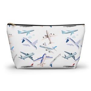 May include: White cosmetic bag with a black zipper and gold pull. The bag is covered in a pattern of various cartoon airplanes in shades of blue, gray, and red. The planes are depicted in flight against a white background.