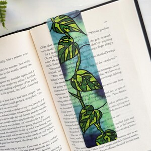 May include: A rectangular bookmark featuring a watercolor-style illustration of a trailing pothos plant with variegated green and yellow leaves. The background consists of blended shades of blue and purple, placed inside an open hardcover book.