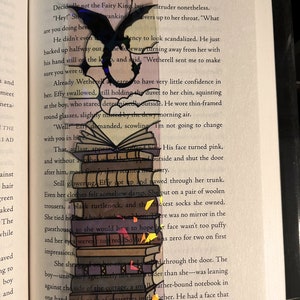 May include: A colorful bookmark featuring a stack of books with a black dragon silhouette. The bookmark is made of clear plastic with a rainbow-colored design.