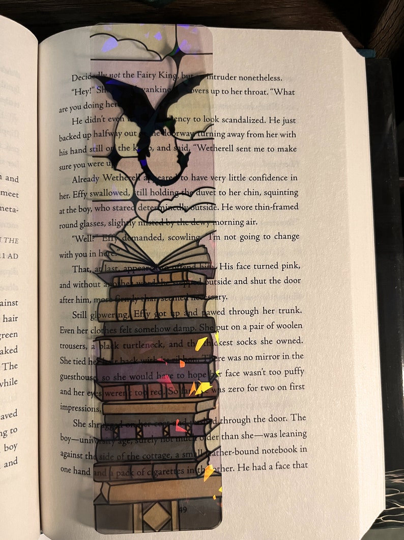 May include: A colorful bookmark featuring a stack of books with a black dragon silhouette. The bookmark is made of clear plastic with a rainbow-colored design.