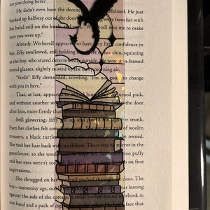 May include: A black and white bookmark with a dragon design. The bookmark is layered with a stack of books and has a pink and purple background.