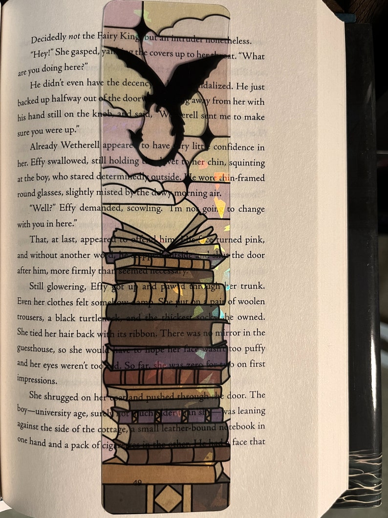 May include: A black and white bookmark with a dragon design. The bookmark is layered with a stack of books and has a pink and purple background.