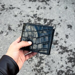 Puede incluir: Una cartera negra de dos pliegues con un patrón texturizado similar al de un cocodrilo. La cartera se sostiene en una mano, mostrando su acabado brillante. La cartera parece ser de cuero.