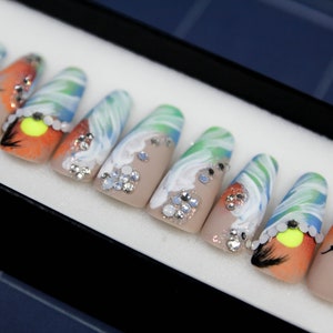 May include: A set of ten false nails with a beach-themed design. The nails are painted with a light brown base and feature a variety of designs, including waves, sunsets, and seashells. The nails are also decorated with rhinestones.
