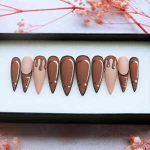 May include: Set of ten false nails with a stiletto shape. The nails are painted in a light brown colour with a brown drip design on one nail.