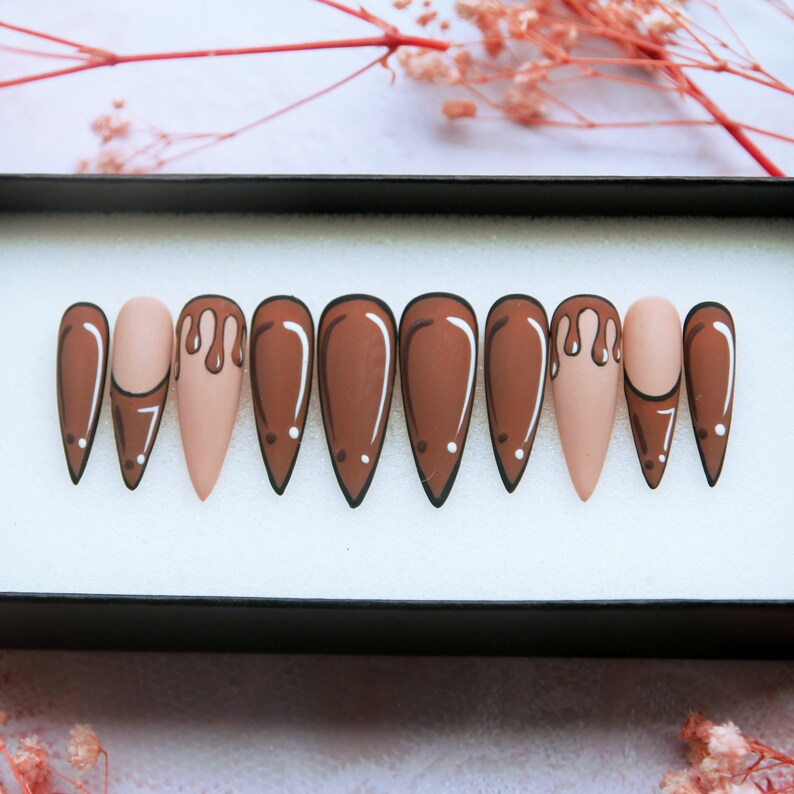 May include: Set of ten false nails with a stiletto shape. The nails are painted in a light brown colour with a brown drip design on one nail.