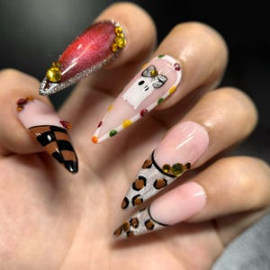 Könnte beinhalten: Stiletto-förmige künstliche Fingernägel mit verschiedenen Designs, darunter ein rotes Katzenaugen-Finish, ein weißes Gespenst mit Schleife, schwarz-braune Karomuster und Leopardenmuster, verziert mit kleinen bunten Strasssteinen.
