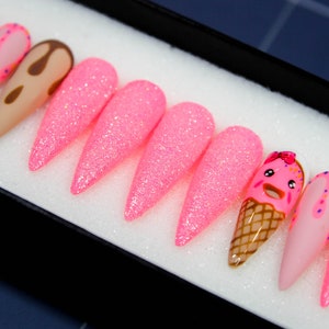 May include: Set of ten pink glitter press-on nails with various designs, including a brown chocolate drip, a pink ice cream cone with a smiling face, and a pink nail with white and blue sprinkles.