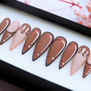 May include: Set of ten false nails with a stiletto shape. The nails are painted in a light brown colour with a white outline. Some of the nails have a brown drip design.