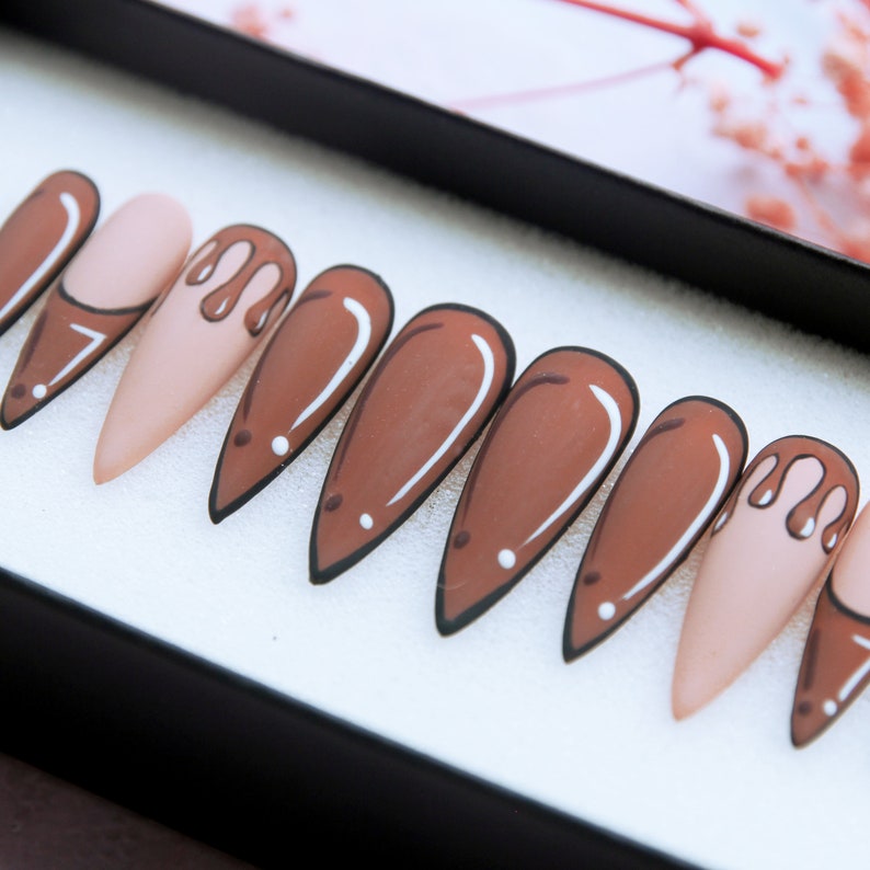 May include: Set of ten false nails with a stiletto shape. The nails are painted in a light brown colour with a white outline. Some of the nails have a brown drip design.
