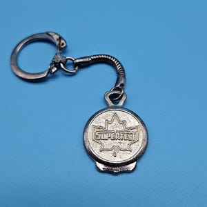May include: Silver-coloured keychain with a circular pendant featuring a maple leaf design and the word "SUPERTEST". The keychain has a ring and a flexible, coiled connecting piece. The background is a solid blue.