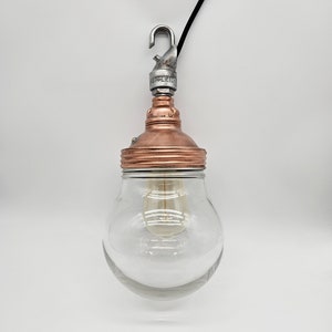 May include: A copper and glass pendant light with a clear glass shade and a silver hook. The light is hanging from a black cord.