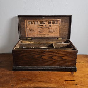 Op de afbeelding: Een open, antieke houten gereedschapskist met de tekst "BOYS 'LITTLE DAISY' TOOL CHEST" op de binnenkant van het deksel. De kist is donkerbruin met zichtbare houtnerf en bevat kleine compartimenten en gereedschap. De kist staat op een houten oppervlak.