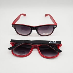 May include: Red and black sunglasses with a Coca-Cola logo on the side. The sunglasses have a UV protection label.