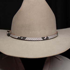 May include: A light beige cowboy hat featuring a narrow hatband with a textured snakeskin pattern in alternating black and white scales. The hatband includes a small, rectangular metal buckle on the left side, securing the band around the crown.