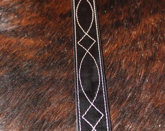 May include: Close-up of a black leather belt with white stitching. The stitching forms a repeating pattern of diamonds and elongated ovals. The belt is set against a brown and black textured background.