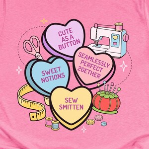 May include: Pink t-shirt featuring a sewing-themed design. The design includes heart-shaped candies with phrases like "Cute as a Button" and "Sew Smitten", along with a sewing machine, scissors, and measuring tape. A fun and creative gift for a seamstress.