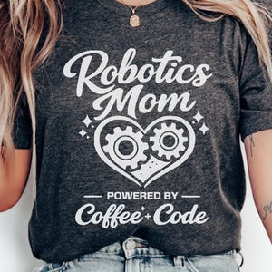 May include: Dark gray t-shirt with the text "Robotics Mom" above a heart-shaped design with gears. Below the heart, the text reads "Powered by Coffee + Code". The shirt is paired with light blue jeans and white sneakers.
