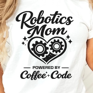 May include: White t-shirt with the text "Robotics Mom" above a heart-shaped design with gears and the words "Powered by Coffee + Code". The shirt is worn by a person in blue jeans.
