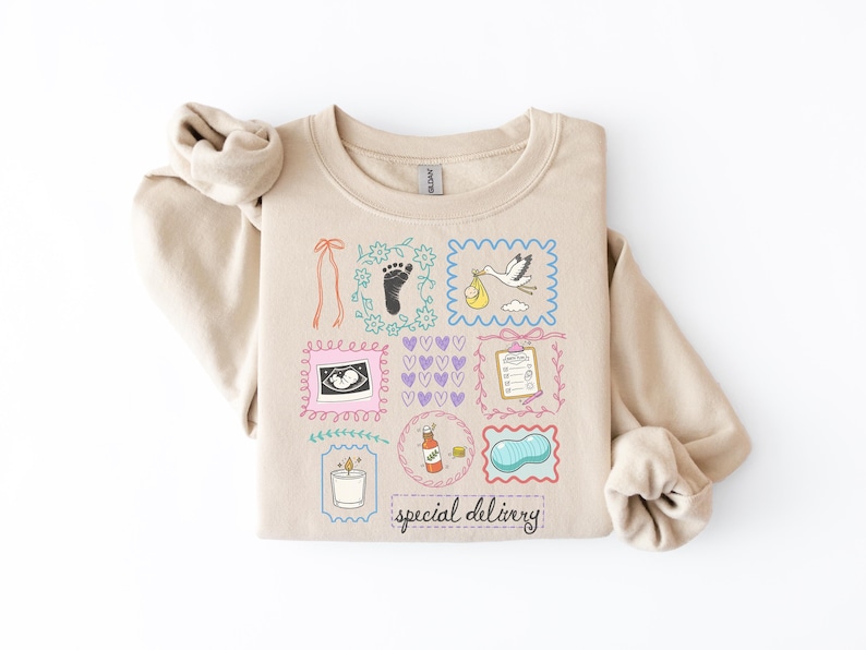 Peut inclure: Sweat-shirt beige avec un motif graphique color&eacute; repr&eacute;sentant des illustrations li&eacute;es aux b&eacute;b&eacute;s. Le motif comprend une cigogne portant un b&eacute;b&eacute; et les mots "special delivery".