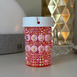 Puede incluir: Un frasco de pastillas rosa cubierto de pedrería con las palabras "SEXY LEXY" escritas con cuentas de letras doradas. El frasco tiene una tapa blanca y está decorado con varios tonos de pedrería y cuentas rosas.