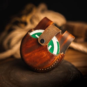 May include: A handcrafted, brown leather coin purse with a flap secured by a black snap. The purse holds several green and white items. The leather has a distressed appearance, and the purse is set on a dark wooden surface.