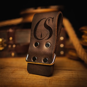 May include: A brown leather belt loop with a gold metal buckle and the initials "CS" engraved on the front.