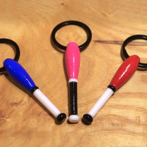 Puede incluir: Tres clavas de malabares de colores con anillos negros y puntas blancas. Las clavas son roja, azul y rosa.