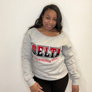 May include: Gray sweatshirt with a red and black "DELTA" graphic and the text "Devastating Diva" underneath.
