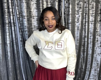 May include: A white sweatshirt with a gold and maroon Greek letter design that says "ΔΣΘ" and "1913" on the sleeve. The sweatshirt is worn by a person with a maroon skirt.
