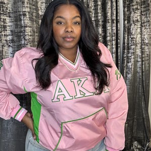 Puede incluir: Chaqueta rosa y verde con las letras "AKA" bordadas en la parte delantera. La chaqueta tiene cuello en V, un bolsillo delantero y puños a rayas verdes y blancas. La chaqueta la lleva una persona con pelo largo y oscuro.