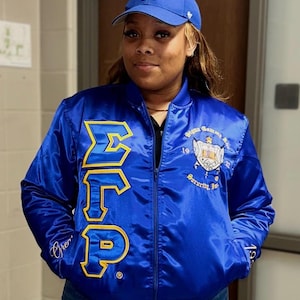 Könnte beinhalten: Blaue Satin-Bomberjacke mit goldfarbenen Buchstaben und Aufnähern. Die Jacke zeigt die griechischen Buchstaben ΣΓΡ in Gold, zusammen mit einem Wappen und den Worten "Sigma Gamma Rho Sorority, Inc." Eine blaue Baseballkappe mit weißem "LA"-Logo ist ebenfalls zu sehen.