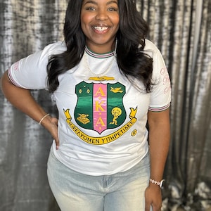 May include: White t-shirt with a crest design featuring the letters AKA in pink and gold, with green and yellow accents. The shirt has pink and green striped trim on the sleeves and collar. The shirt also has text on the sleeves.
