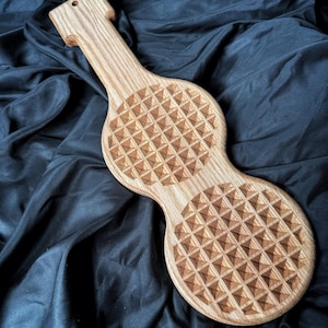 May include: A light brown wooden paddle featuring a long handle with a small hole at the top and two circular sections. Each circular section is engraved with a repeating geometric pattern of small, raised pyramids across the entire surface.