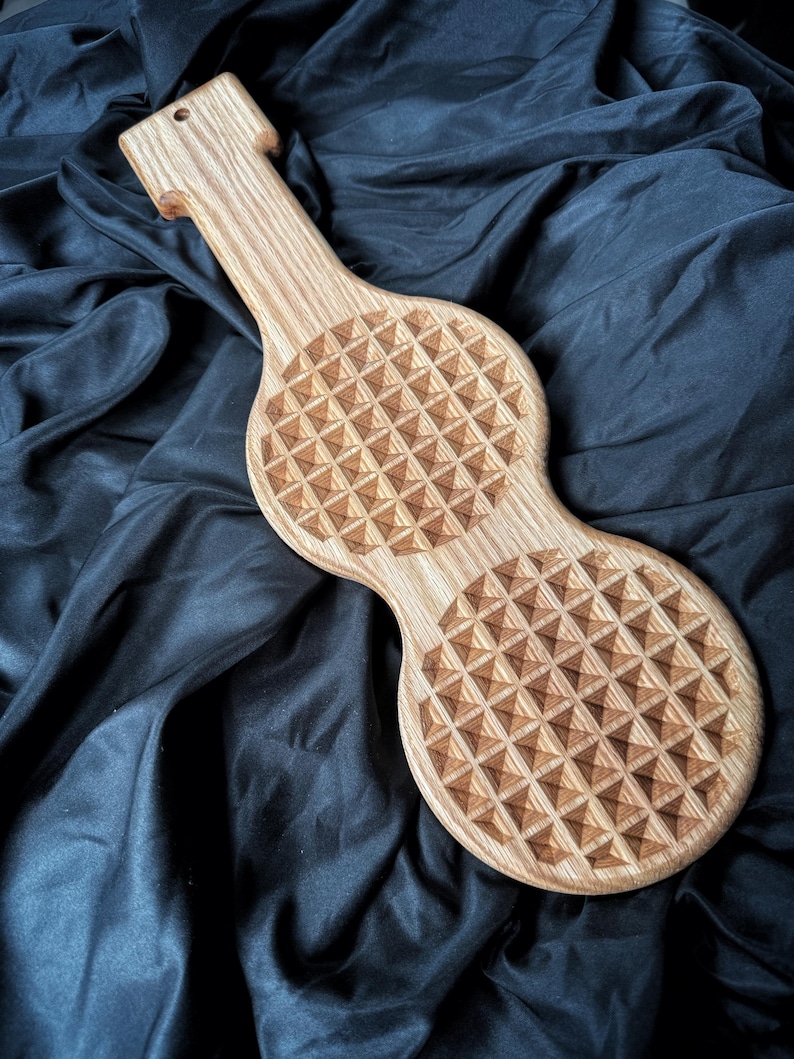 May include: A light brown wooden paddle featuring a long handle with a small hole at the top and two circular sections. Each circular section is engraved with a repeating geometric pattern of small, raised pyramids across the entire surface.