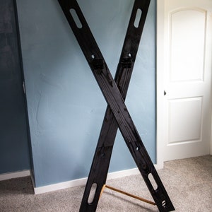 May include: A black wooden X-shaped stand with two wooden dowels. The stand is on a light brown carpet.