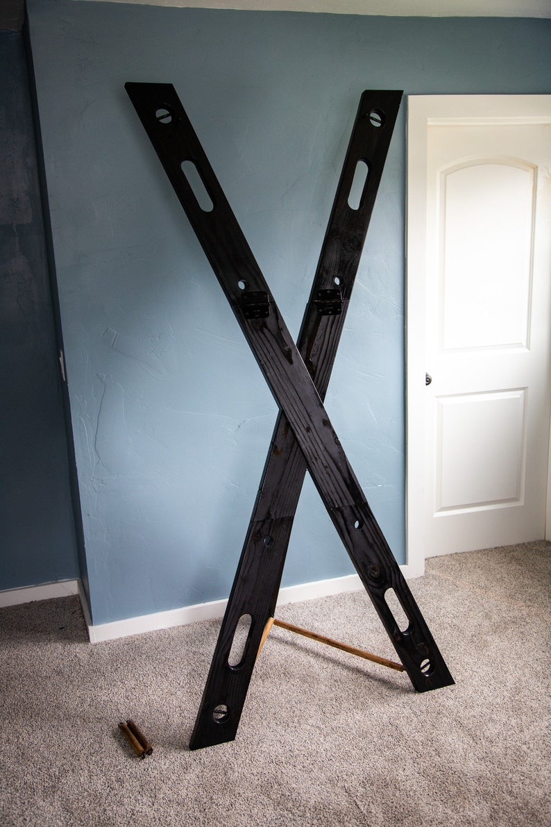 May include: A black wooden X-shaped stand with two wooden dowels. The stand is on a light brown carpet.
