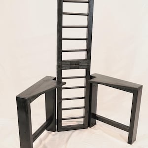 Puede incluir: Una estructura de madera negra que presenta un panel vertical alto similar a una escalera, conectado mediante bisagras met&aacute;licas a dos plataformas laterales triangulares. La pieza est&aacute; hecha de madera te&ntilde;ida oscura con veta visible.