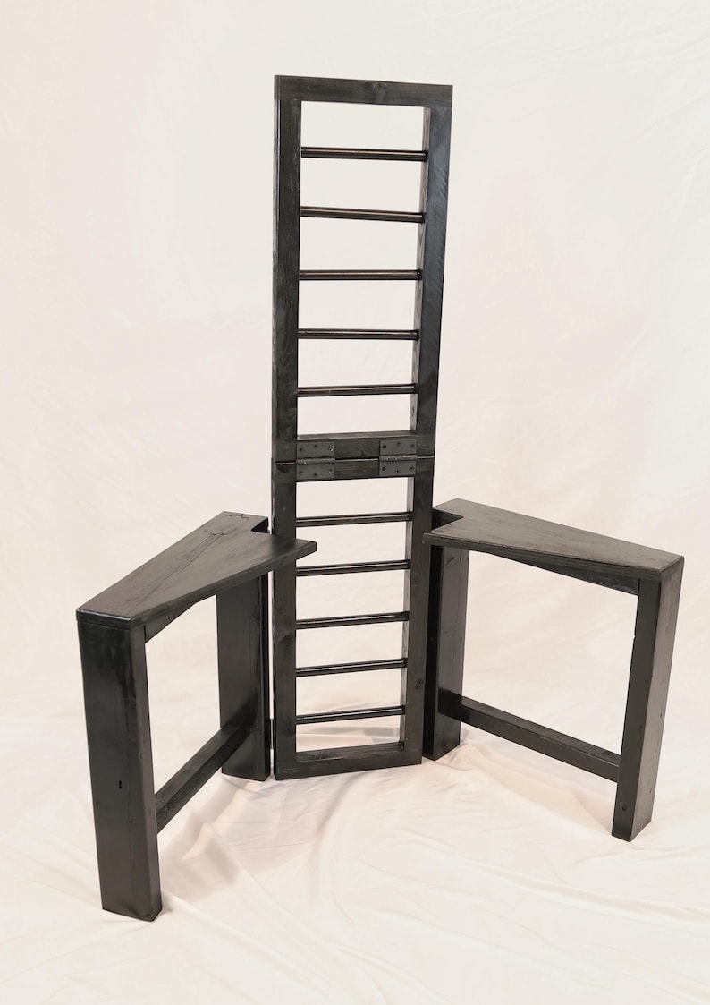 Puede incluir: Una estructura de madera negra que presenta un panel vertical alto similar a una escalera, conectado mediante bisagras met&aacute;licas a dos plataformas laterales triangulares. La pieza est&aacute; hecha de madera te&ntilde;ida oscura con veta visible.