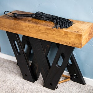 May include: A wooden table with a black, X-shaped base. The table top is made of a single piece of wood and has a natural finish. A black leather whip is lying on the table top.