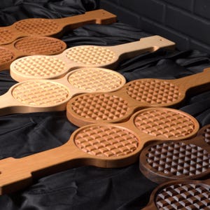 May include: A collection of wooden kitchen tools, each featuring a long handle and two circular heads with a raised, grid-like pyramid texture. The tools are crafted from various wood tones, ranging from light maple to dark walnut, arranged in a group.