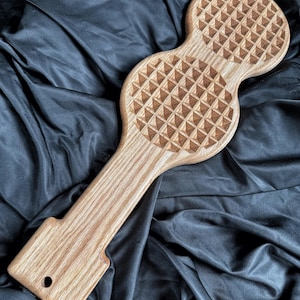 May include: A light brown wooden kitchen tool featuring a long handle and two circular heads with a raised, grid-like pyramid texture. The handle includes a small circular hole at the base for hanging, showcasing a natural wood grain pattern.