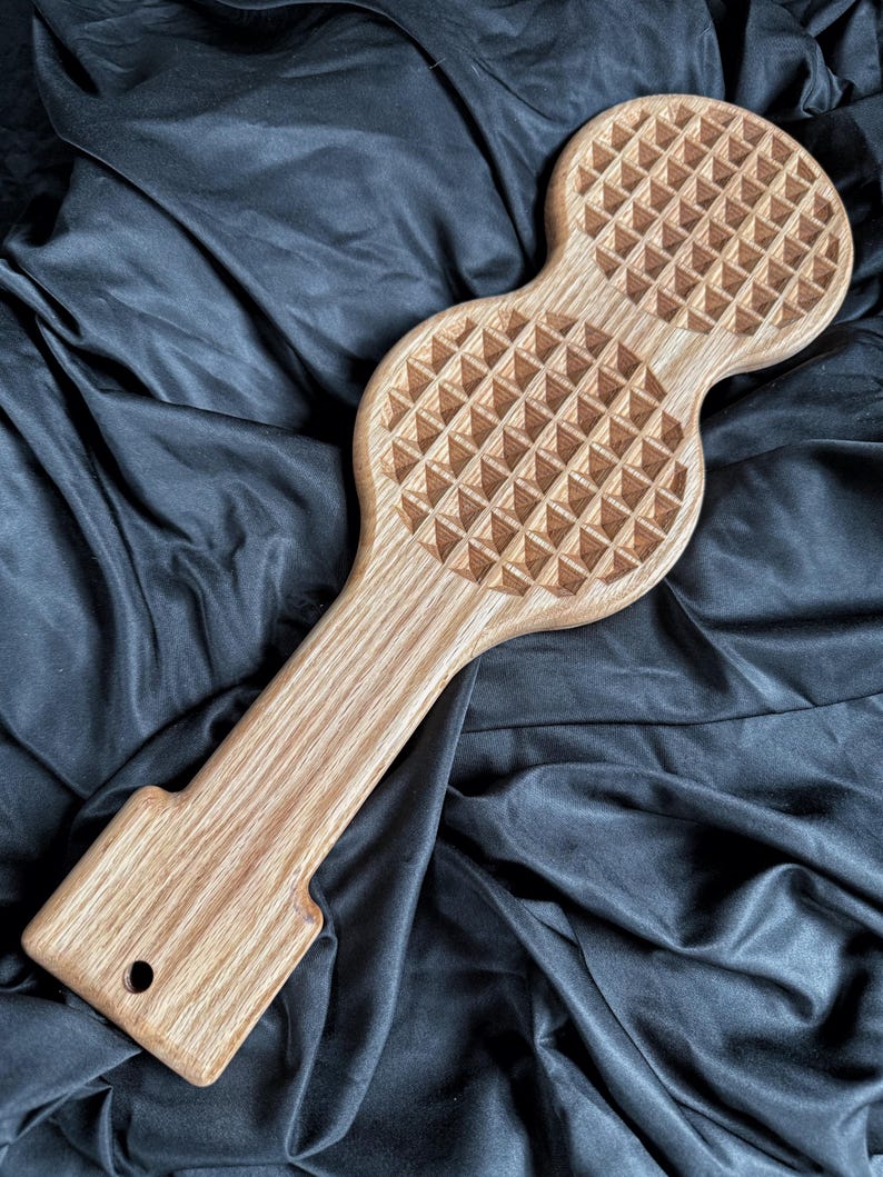 May include: A light brown wooden kitchen tool featuring a long handle and two circular heads with a raised, grid-like pyramid texture. The handle includes a small circular hole at the base for hanging, showcasing a natural wood grain pattern.