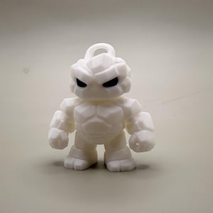 May include: A white figurine of a stylized, muscular character with a rocky texture. The figure has black eyes and a loop at the top, suggesting it could be a keychain or ornament. The design is reminiscent of a superhero or fantasy character.