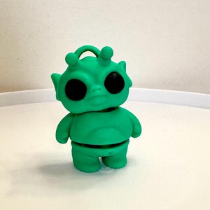 May include: A green alien figurine with large black eyes, small arms, and a loop on top. The alien has a segmented body and is standing on a white surface. The figurine is a solid, bright green color.