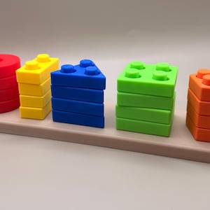 May include: A colorful wooden stacking toy featuring five different shapes: a red cylinder, yellow rectangles, a blue triangle, a green square, and an orange hexagon. The shapes are stacked on a light brown rectangular base. Educational toy for toddlers.