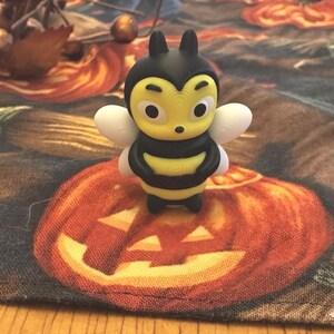 May include: A 3D-printed bee figurine with a yellow body, black stripes, and white wings. The bee has large eyes and is standing on a Halloween-themed fabric with pumpkins.