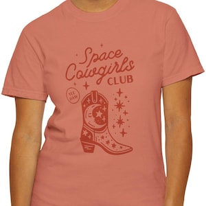 May include: A coral colored t-shirt with the text "Space Cowgirls Club" and a stylized illustration of a cowboy boot with a crescent moon and stars.